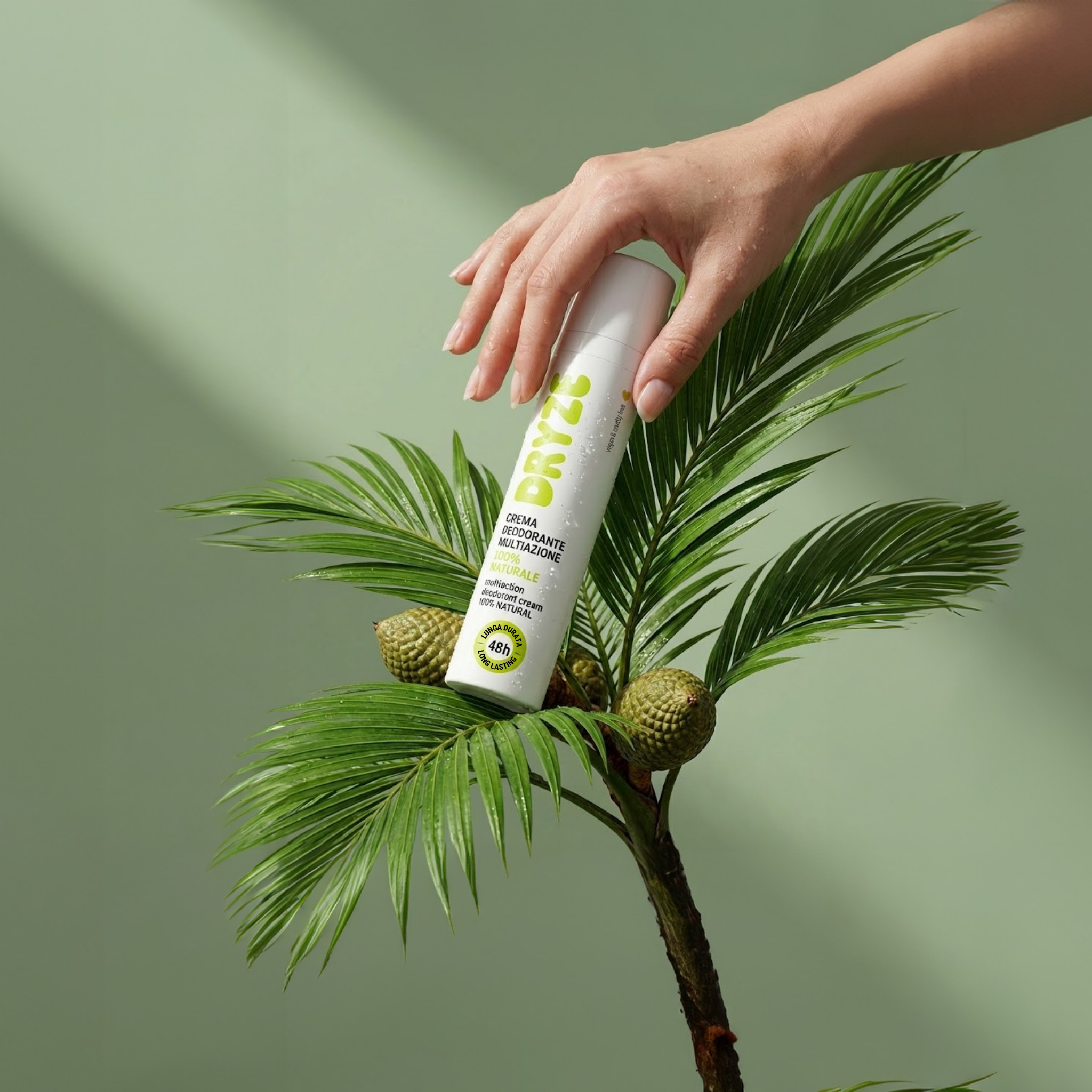 Mano che regge il flacone di Dryze Crema Deodorante Multiazione tra foglie di palma su sfondo verde, formato verticale per mobile.